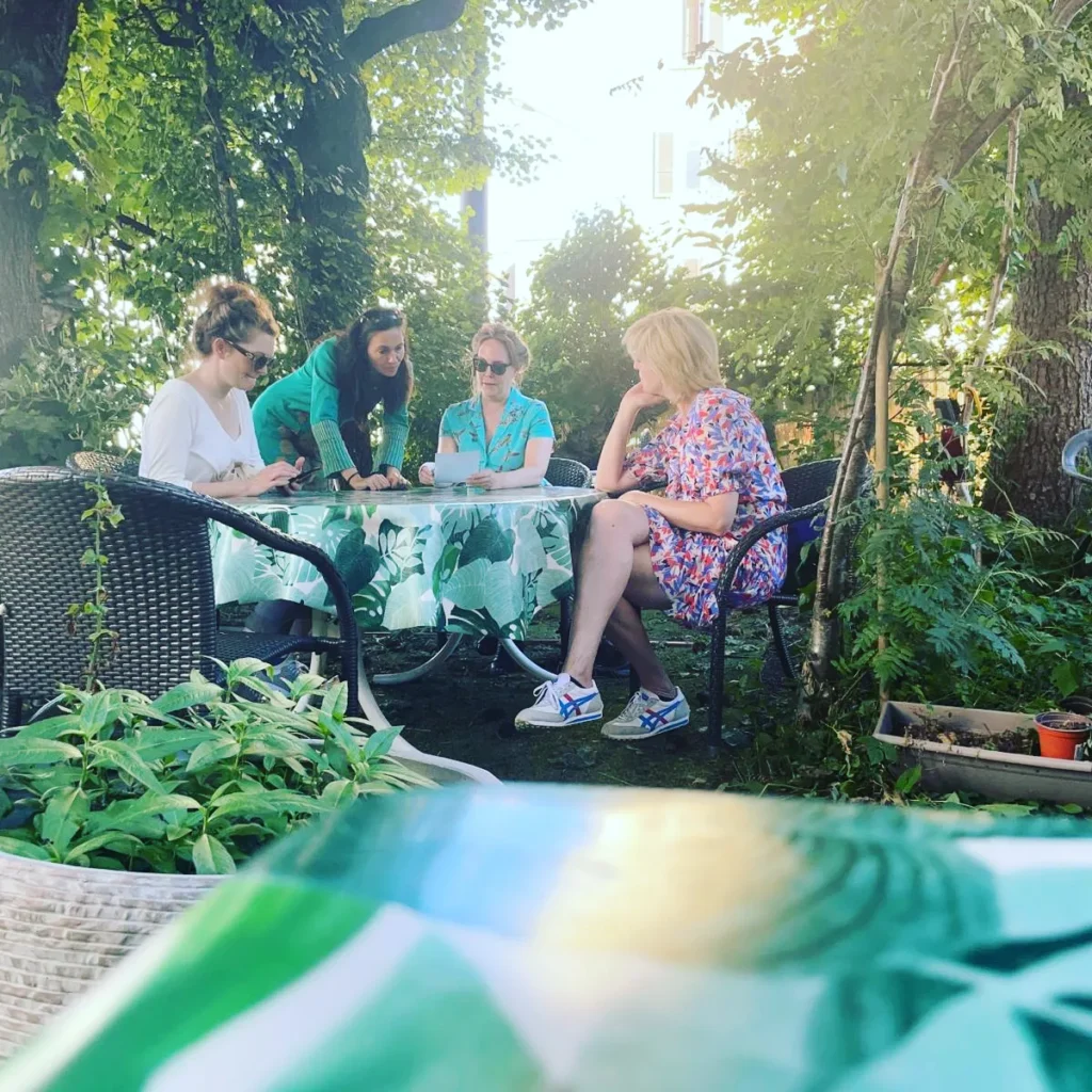Ein Gruppenfoto im Grünen, 4 Personen sitzen um einen Gartentisch, runherum sind grüne Pflanzen. Drei Personen unterhalten sich, die vierte ist mit ihrem Smartphone beschäftigt.